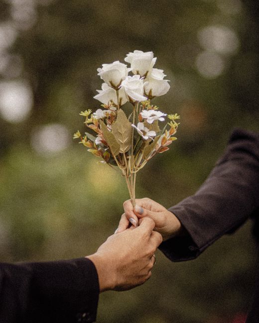A person in a dark suit is handing over a bouquet of white roses with green foliage and small light pink accent flowers, wrapped loosely with natural paper and tied with a simple ribbon, in an outdoor