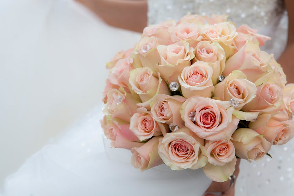 A close-up of a floral bouquet featuring numerous pale pink roses with soft, velvety petals arranged in a compact, rounded style. The roses are accented with small, sparkling rhinestones placed among 