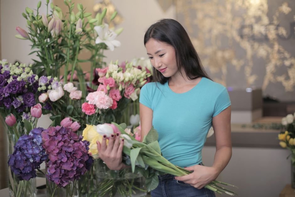 A young woman with long black hair and light skin, wearing a light blue t-shirt, is holding a bouquet of mixed flowers, including white tulips and green foliage, in a flower shop. She is standing besi