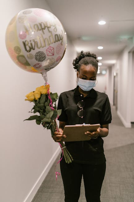 A woman wearing a light blue face mask and black uniform stands in a well-lit corridor, holding a bouquet of fresh yellow roses wrapped in dark green foliage and pink ribbon. In her other hand, she ho