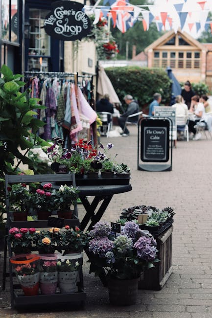 An outdoor flower display outside The Green's Cut Shop on a busy street near Clitterhouse Playing Fields, featuring vibrant potted flowers including purple hydrangeas, pink and red petunias, and other