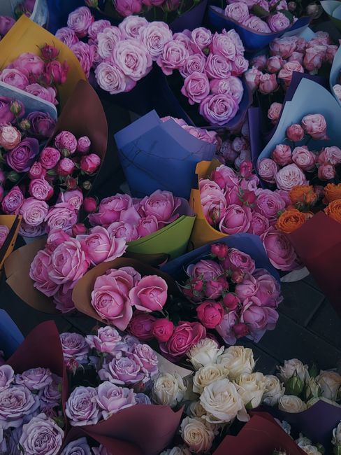 A vibrant assortment of freshly arranged flower bouquets featuring pink, purple, white, and orange roses, along with peonies and ranunculus. The flowers are wrapped in colorful paper in shades of blue